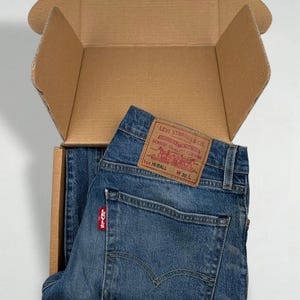 Könnte beinhalten: Blaue Jeans aus Denim, gefaltet in einem Versandkarton aus Pappe. Die Jeans hat einen beigen Lederaufnäher am hinteren Bund mit dem Text Levi Strauss & Co. Genuine Quality Clothing, Lot Hi-Ball, W 30 L und ein rotes Levi's-Etikett.