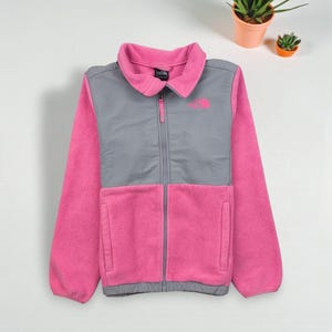 May include: A pink and gray fleece jacket with a full zipper closure. The jacket features a gray chest panel and pink sleeves and body. The North Face logo is visible on the chest. Two front pockets are also visible.