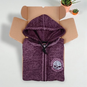 May include: A purple zip-up hoodie with a Harley-Davidson skull logo, displayed inside an open cardboard box. The hoodie has a drawstring hood and a full-length zipper. Small potted plants are in the background.