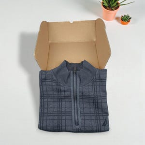 Puede incluir: Una camisa gris a cuadros con cremallera está cuidadosamente doblada dentro de una caja de cartón abierta. La camisa tiene un cuello alto y una cremallera plateada. Una pequeña planta en maceta y una suculenta están en el fondo.