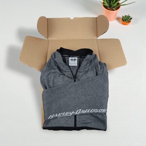 May include: A gray Harley-Davidson zip-up jacket is neatly folded inside an open cardboard box. The jacket has a black collar and the Harley-Davidson logo is visible. Two small potted plants are in the background.