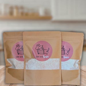 May include: Three brown kraft paper stand-up pouches with clear windows containing a white granular substance. Each pouch features a circular pink label with a black line drawing of a dachshund wearing a beret and sweater, holding an envelope, with the text "LENNY WITH love".