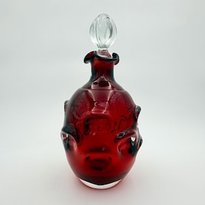 May include: A deep red glass decanter featuring a bulbous body with irregular indentations and a dark green tint near the rim. The vessel is topped with a clear, spherical glass stopper that has a subtle ribbed texture on its surface.