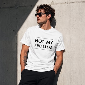 Könnte beinhalten: Ein weißes kurzärmeliges T-Shirt mit dem schwarzen Text NOT MY PROBLEM, der mittig auf der Brust zwischen zwei horizontalen schwarzen Linien platziert ist. Das T-Shirt hat einen Rundhalsausschnitt und eine normale Passform, kombiniert mit einer schwarzen Hose und einer dunklen Sonnenbrille.