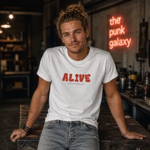 Könnte beinhalten: Weißes T-Shirt mit dem Wort "ALIVE" in Rot und "THEPUNKGALXY" darunter. Das T-Shirt hat kurze Ärmel und einen Rundhalsausschnitt. Im Hintergrund befindet sich ein Neonschild mit der Aufschrift "the punk galaxy".
