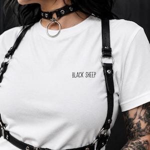 Könnte beinhalten: Ein weißes T-Shirt mit dem schwarzen Aufdruck BLACK SHEEP auf der Brust. Das T-Shirt wird mit einem schwarzen Ledergeschirr kombiniert, das silberfarbene Metallringe, Nieten und verstellbare Riemen sowie ein passendes Lederhalsband aufweist.