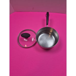 May include: A stainless steel saucepan with a black handle and a clear glass lid with a black knob. The lid is off to the side, revealing the interior of the pan. The pan is on a pink surface.