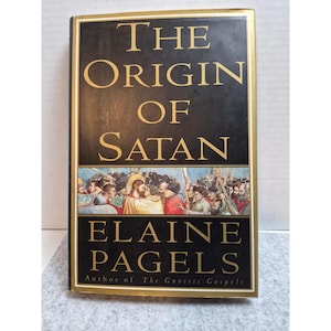 May include: A hardcover book titled The Origin of Satan by Elaine Pagels. The black cover features gold lettering and a central horizontal illustration depicting a historical scene with figures in robes, including a central haloed person.