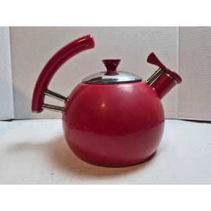 May include: A round, bright red tea kettle with a glossy finish. It features a curved red handle attached with two silver metal rods, a silver metal lid with a red knob, and a red spout with a silver metal tip and a small red lever mechanism.