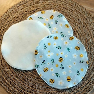 Könnte beinhalten: Drei runde, wiederverwendbare Abschminkpads. Zwei Pads haben einen hellblauen Hintergrund mit einem Muster aus Bienen, weißen Gänseblümchen und grünen Blättern. Das dritte Pad ist schlicht weiß. Die Pads liegen auf einem gewebten, braunen Platzset.