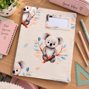 Può includere: Una copertina rettangolare in tessuto per raccoglitore con un motivo ripetuto di koala illustrati appollaiati su rami arancioni con foglie blu e arancioni. La copertina include una finestra rettangolare in plastica trasparente per un'etichetta.