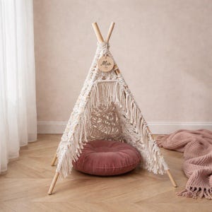 Op de afbeelding: Een crèmekleurige macramé tipi-tent met houten palen en een rond, stoffig roze kussen binnenin. Een houten label met de naam "Mia" hangt bovenaan. Een bijpassende deken is in de buurt gedrapeerd. De tent is voor kinderen.