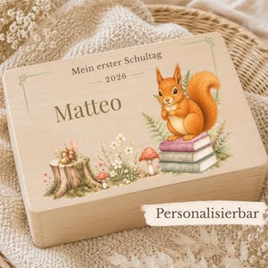 Könnte beinhalten: Eine rechteckige Holzbox mit einem aufgedruckten Bild eines Eichhörnchens auf einem Bücherstapel, einem Baumstumpf mit Eicheln und Pilzen. Der Text lautet "Mein erster Schultag", "2026", "Matteo" sowie "Personalisierbar".