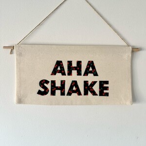 May include: A white canvas banner with black text that reads "AHA SHAKE" with a cherry pattern on the letters. The banner is hanging from a string.