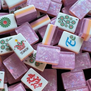 May include: A collection of mahjong tiles featuring white faces with green floral patterns, a red dragon, a blue letter E with a flower, and a colorful joker tile with the text JOKER. The tiles have translucent, iridescent pink backs.