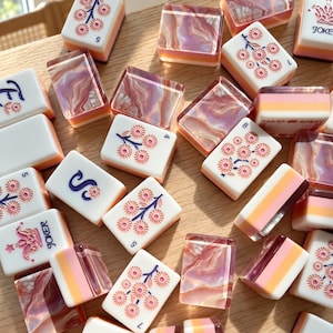 Puede incluir: Una colección de fichas de mahjong rectangulares con caras blancas, motivos florales rosas, letras azules y etiquetas Joker. Algunas fichas tienen dorsos translúcidos de color rosa y naranja con efecto mármol.