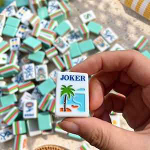 May include: A rectangular white Mahjong tile featuring the word JOKER in blue text above a graphic of a green palm tree on a sandy beach with blue water. The tile is held against a background of numerous other colorful Mahjong tiles.