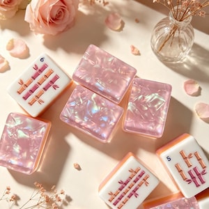 May include: A collection of rectangular mahjong tiles featuring a glossy, iridescent pink finish with a pearlescent texture. Some tiles display bamboo patterns in pink and brown with the numbers 5 or 6 printed in blue on the white surface.