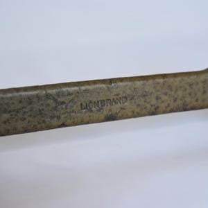 Op de afbeelding: Een metalen moersleutel in vintage-stijl met een textuurrijke, gevlekte bronskleurige afwerking. De centrale schacht is voorzien van de in reliëf aangebrachte tekst LIONBRAND in hoofdletters. Het gereedschap heeft aan beide zijden open uiteinden.