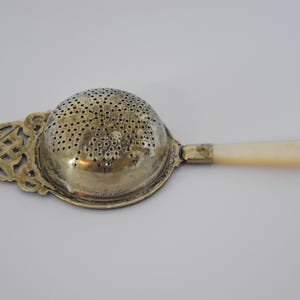 May include: A silver-toned metal tea strainer featuring a perforated dome-shaped bowl, an ornate filigree handle attachment with geometric cutouts, and a smooth, cylindrical off-white handle made of a polished, opaque material.
