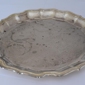 Puede incluir: Una bandeja de servicio redonda de metal plateado con un borde festoneado y ondulado. La superficie de la bandeja presenta un acabado reflectante con manchas oscuras visibles, deslustre y marcas circulares en el centro.
