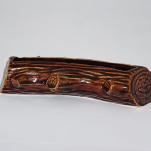 May include: A brown ceramic planter shaped like a hollowed-out log. The exterior features a glossy finish with molded bark texture, including knots and a circular tree ring pattern on the end. The top has a long, narrow opening for plants.