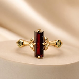 Peut inclure: Une bague en or avec une pierre précieuse grenat rouge foncé taille baguette verticale, flanquée de deux petites pierres vertes taille ronde. L'anneau est orné de motifs de feuilles dorées de chaque côté du sertissage central.