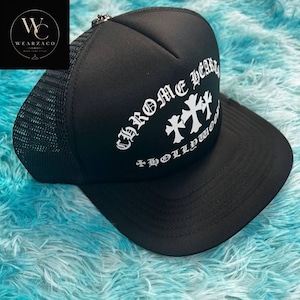 Puede incluir: Gorra de camionero negra con parte trasera de malla y visera curva. La parte delantera de la gorra presenta el texto "Chrome Hearts Hollywood" en blanco, junto con dos diseños de cruces blancas. Cierre a presión.