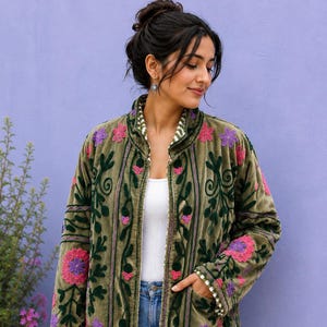 Puede incluir: Una chaqueta de terciopelo verde oliva con mangas largas, cuello alzado y bolsillos delanteros. La chaqueta está decorada con motivos florales bordados en tonos rosa, morado y verde oscuro, con un forro interior a rayas blancas y verdes.