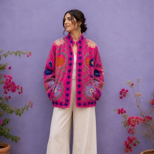 Könnte beinhalten: Eine leuchtend fuchsiafarbene Jacke mit aufwendiger, mehrfarbiger Blumenstickerei, Stehkragen und langen Ärmeln, kombiniert mit einer weiten, cremeweißen Hose und schlichten braunen flachen Schuhen. Die Jacke hat einen Knopfverschluss vorne.