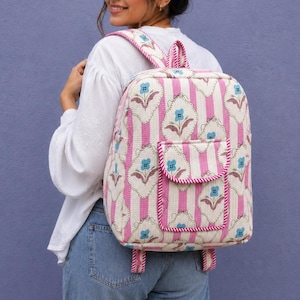 Puede incluir: Una mochila con un patrón de rayas verticales rosas y blancas y un motivo floral repetitivo de flores azules sobre formas festoneadas de color crema. La bolsa incluye un bolsillo frontal con cierre de solapa y dos correas ajustables.