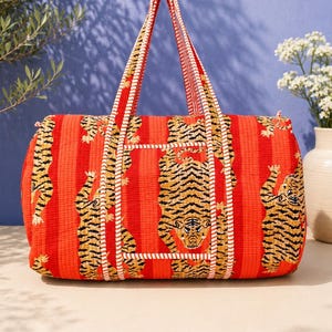 Könnte beinhalten: Eine rechteckige, gesteppte Reisetasche in leuchtendem Orangerot mit einem sich wiederholenden Muster aus schwarzen und beigen Tigern. Die Tasche verfügt über zwei lange Tragegriffe mit weiß-roter Streifenverzierung, die auch die Struktur betont.