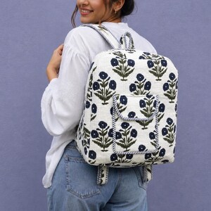 Puede incluir: Una mochila blanca con un patrón floral repetitivo en azul oscuro y verde. La bolsa tiene forma rectangular, un asa superior, dos correas para los hombros y un bolsillo frontal con cierre de solapa. La tela tiene un aspecto texturizado y acolchado.