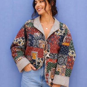 Op de afbeelding: Een gewatteerd jack met een veelkleurig patchworkpatroon met bloemmotieven en geometrische vormen in tinten rood, marineblauw, groen en mosterdgeel. Het jack heeft een omgeslagen kraag en brede manchetten met een wit en bruin gestreepte voering.