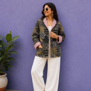 Könnte beinhalten: Eine langärmelige Jacke mit dunkler Basis und buntem abstraktem Muster, einem Reverskragen mit roten und weißen Streifen sowie passenden gestreiften Ärmelbündchen. Die Jacke wird über einem schlichten weißen Oberteil und einer weißen weiten Hose getragen.