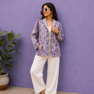 May include: A long-sleeve jacket featuring a purple and white intricate floral pattern with a blue and white striped collar lining, worn over a white jumpsuit. The jacket has two visible buttons on the front and two side pockets.
