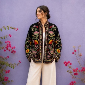 Könnte beinhalten: Eine schwarze Jacke mit langen Ärmeln und Stehkragen, verziert mit farbenfrohen Blumenstickereien in Rosa, Gelb und Grün. Die Jacke wird über einem weißen Oberteil und einer weißen Hose mit weitem Bein getragen, dazu braune flache Schuhe.