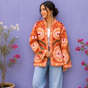 Könnte beinhalten: Eine orangefarbene Langarmjacke mit aufwendigen Blumenstickereien in Rosa, Weiß und Schwarz. Die Jacke hat einen Stehkragen und eine offene Vorderseite, getragen über einem weißen Oberteil mit hellblauer, weit geschnittener Jeans und beigen Sandalen.