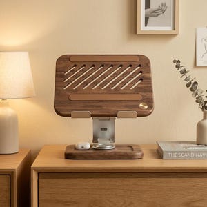 Peut inclure: Un support pour ordinateur portable composé d'une plateforme en bois de noyer brun foncé avec des fentes de ventilation diagonales et deux bandes de préhension en silicone brun. Il comprend un bras de support réglable en métal argenté et une base.