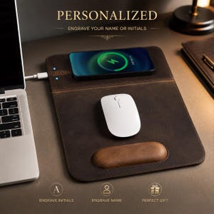 Pode incluir: Um tapete de secretária em pele castanha escura com carregador sem fios integrado para smartphone, um rato de computador branco e um apoio de pulso almofadado. O tapete apresenta o texto PERSONALIZED ENGRAVE YOUR NAME OR INITIALS, ENGRAVE INITIALS, ENGRAVE NAME e PERFECT GIFT.