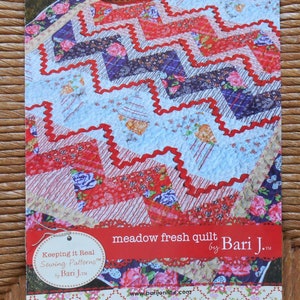 May include: A colorful quilt pattern with a picnic theme. The quilt features a zig-zag design in red, blue, and floral patterns. A picnic basket and a large pink rose are visible. The text reads "meadow fresh quilt by Bari J."