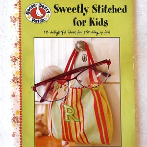 May include: A book titled "Sweetly Stitched for Kids" with a green cover and floral border. A striped bag with the letter "R" and red-framed glasses hang from a hook. The book cover features the Gooseberry Patch logo.