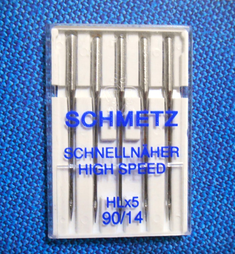 Op de afbeelding: Een verpakking Schmetz-naaimachinenaalden. De naalden zijn zilverkleurig en zitten in een doorzichtige plastic verpakking. De tekst op de verpakking luidt "SCHMETZ SCHNELLN&Auml;HER HIGH SPEED HLx5 90/14". De achtergrond is een blauwe stof.