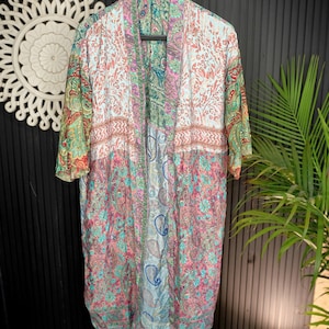 May include: A long, open-front kimono with a vibrant floral and paisley pattern in shades of pink, blue, green, and white. The kimono has short sleeves and is displayed on a black hanger against a dark background.