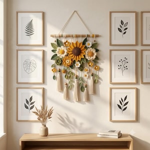 May include: A wall hanging featuring crocheted sunflowers, daisies, and green leaves attached to a wooden dowel with hanging cream-colored tassels and wooden beads. The piece is centered between six framed botanical prints on a light wall.
