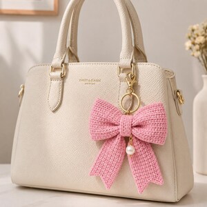 May include: A cream-colored handbag with gold hardware and a pink crocheted bow keychain. The bow has a pearl charm and is attached to a gold keyring. The bag has the text "SWEET & CLASSIC" on the front.