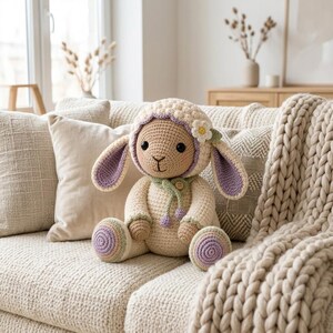 Op de afbeelding: Een gehaakt knuffeldier in de vorm van een lammetje van crèmekleurig garen, met lange paars gevoerde oren, een lichtgroene sjaal met een klein houten knoopje en een witte bloem op het hoofd. De voeten hebben cirkelvormige paarse details.