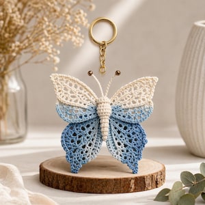 May include: A handmade crochet butterfly keychain in cream and blue hues. The butterfly is displayed on a wooden slice, with a gold-colored keyring attached. The wings transition from cream to shades of blue.