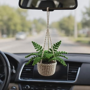 May include: A crochet hanging plant ornament for a car rearview mirror. The piece features a small, beige, textured pot holding a vibrant green fern with detailed, jagged leaves. The plant is suspended by a matching beige braided cord.