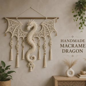 May include: A cream-colored macrame dragon wall hanging suspended from a wooden rod. The dragon features intricate knotted wings, a long coiled tail, and dangling tassels with small beads. The text "HANDMADE MACRAME DRAGON" is visible.